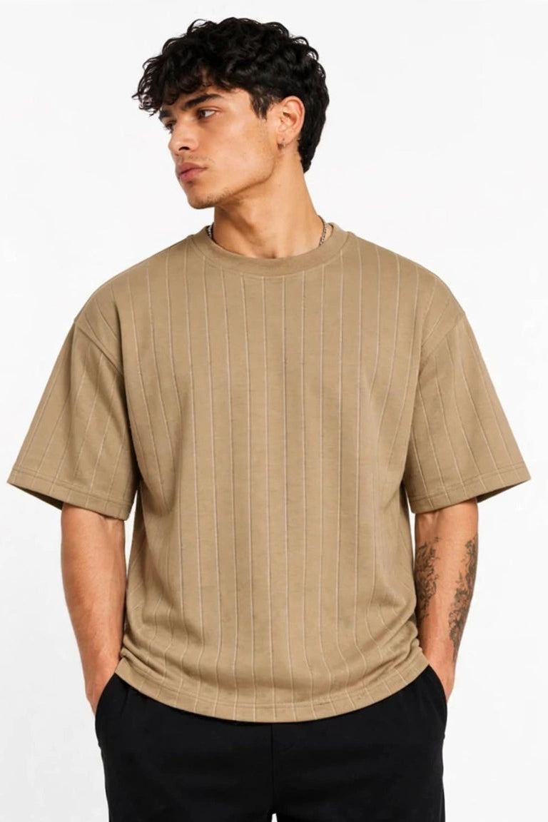 A man wearing a beige short-sleeve crewneck t-shirt with a subtle vertical ribbed texture, paired with black pants, standing against a plain white background.