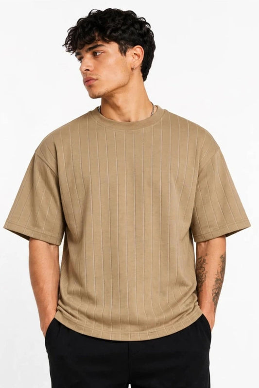 A man wearing a beige short-sleeve crewneck t-shirt with a subtle vertical ribbed texture, paired with black pants, standing against a plain white background.