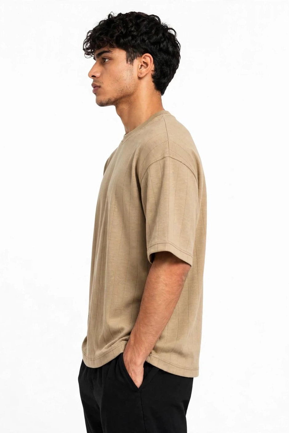 A man in profile wearing a light beige short-sleeve crewneck t-shirt and black pants, standing against a plain white background.