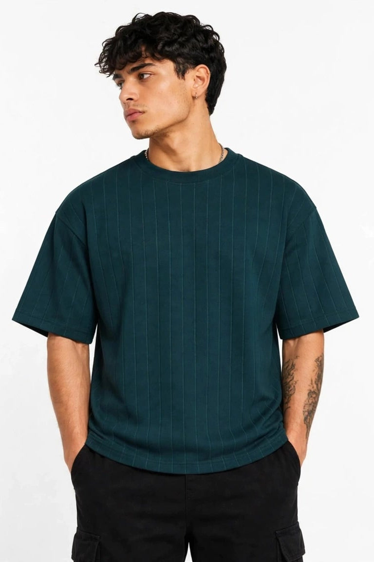 A man wearing a dark teal short-sleeve crewneck t-shirt and black pants, standing against a plain white background.